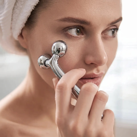 A woman with a white towel wrapped around her head uses a silver facial roller on her cheek.